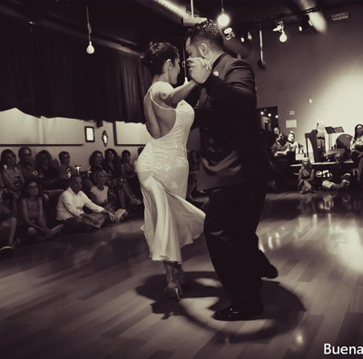 Una coppia balla da vicino in una sala da ballo al ritmo appassionato di Buena Onda Tango, circondata da spettatori seduti. La donna indossa un abito bianco e l'uomo in abiti formali scuri si muove con grazia. La stanza ha un'illuminazione soffusa e un pavimento in legno lucido.
