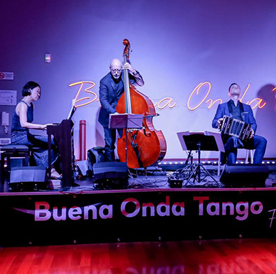 Un trio di musicisti si esibisce su un palco sotto lo slogan "Buena Onda Tango"; uno suona abilmente il pianoforte, l'altro sta in piedi con il contrabbasso e il terzo maneggia magistralmente il bandoneon.