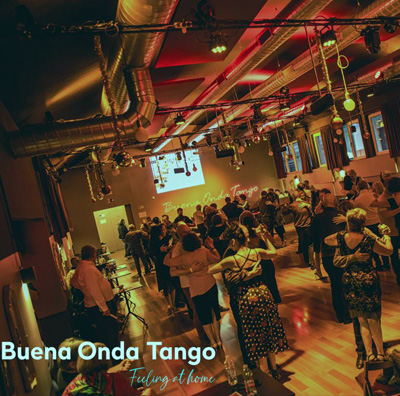 Un gruppo di persone che ballano in una sala da ballo in stile industriale caldamente illuminata con un'insegna proiettata che recita "Buena Onda Tango", trasudando l'energia vibrante e il ritmo dell'autentico Buena Onda Tango.