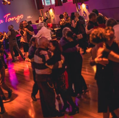 Un gruppo di persone sta ballando il tango Buena Onda in una stanza scarsamente illuminata con pavimenti in legno e insegne al neon sul muro.