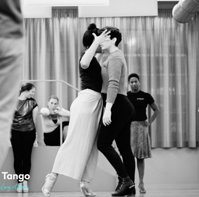 Due individui ballano da vicino in uno studio con altri che osservano in sottofondo, creando un'atmosfera di classe Buena Onda Tango.