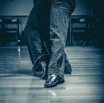Immagine ravvicinata di due persone che ballano insieme su un pavimento di legno; sono visibili solo le gambe e i piedi, che si muovono con grazia in sintonia con i ritmi vivaci di Buena Onda Tango.