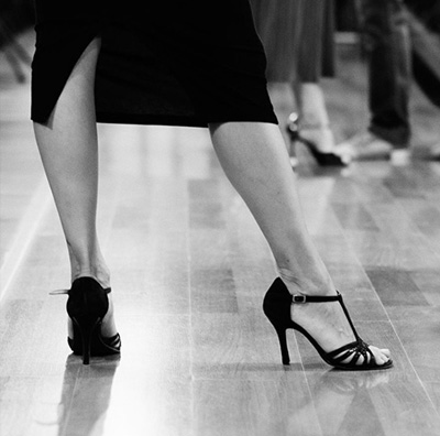 Una persona che indossa scarpe col tacco alto sta con una gamba leggermente piegata, rivelando uno spacco nella gonna, su un pavimento di legno lucido: una vera incarnazione del Buena Onda Tango.