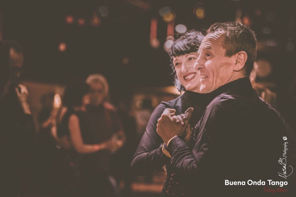 Una donna e un uomo ballano vicini, sorridenti, in un ambiente calorosamente illuminato. Lo sfondo mostra altre persone e alcune luci sfocate. Sono visibili le parole "Buena Onda Tango" e "BelizB Photography".