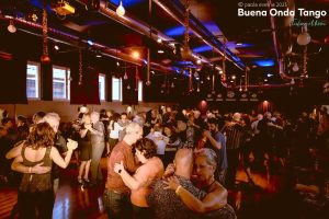 Persone che ballano in coppia durante un affollato evento di tango in una stanza poco illuminata con luci rosse e blu.