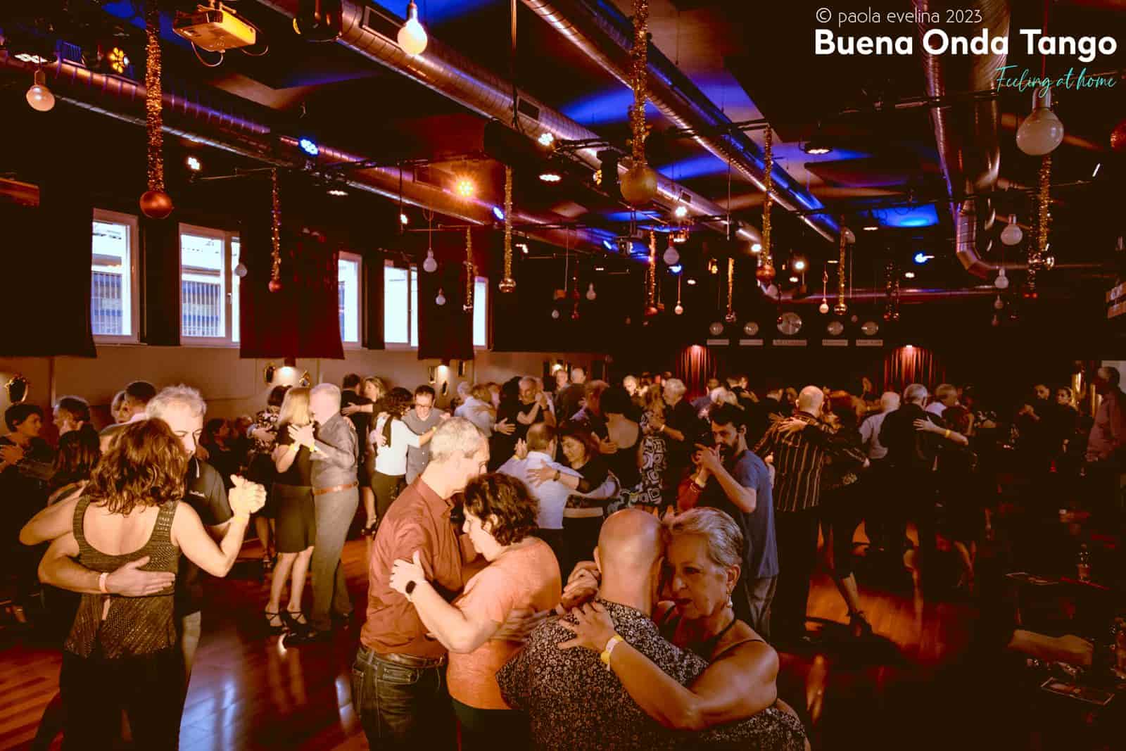 Persone che ballano in coppia durante un affollato evento di tango in una stanza poco illuminata con luci rosse e blu.