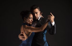 Una coppia balla elegantemente il tango, vestita in abiti formali. La donna indossa un abito blu e l'uomo un completo nero. Sono abbracciati stretti su uno sfondo scuro.
