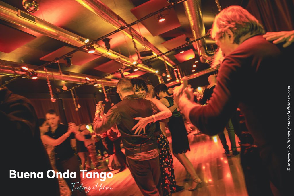 Persone che ballano il tango in una stanza caldamente illuminata con specchi ed elementi metallici sul soffitto. Il testo recita: "Buena Onda Tango - Sentirsi a casa.