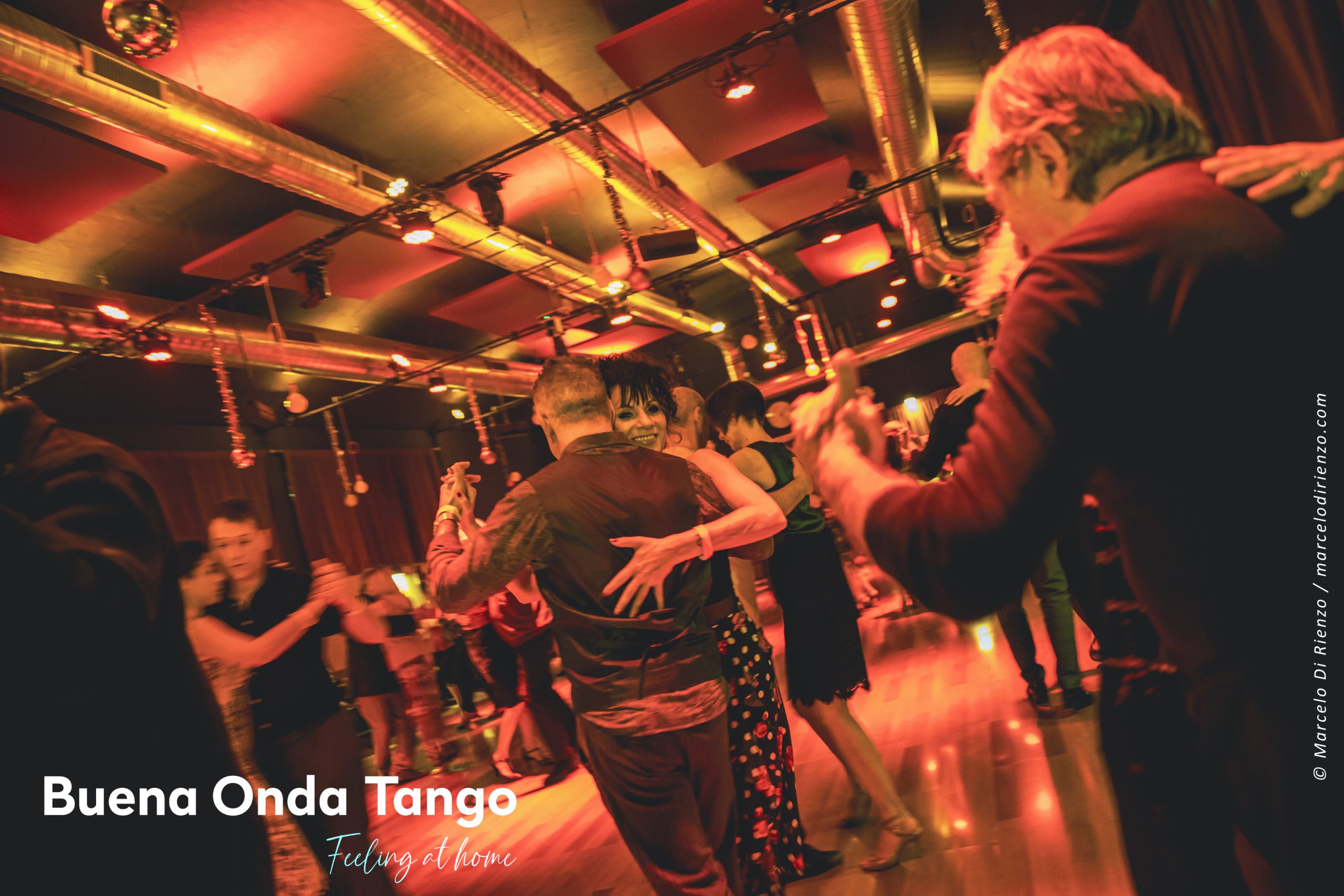 Persone che ballano il tango in una stanza caldamente illuminata con specchi ed elementi metallici sul soffitto. Il testo recita: "Buena Onda Tango - Sentirsi a casa.