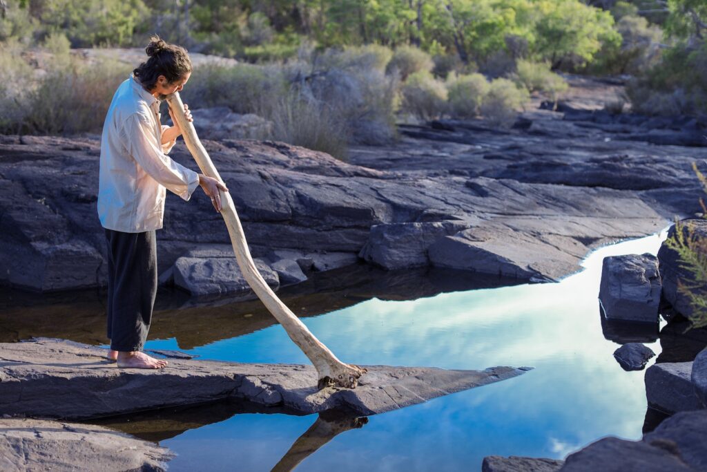 Una persona in piedi a piedi nudi sulle rocce suona un grande didgeridoo vicino a un ruscello poco profondo, nell'acqua del quale si riflettono alberi e cielo.