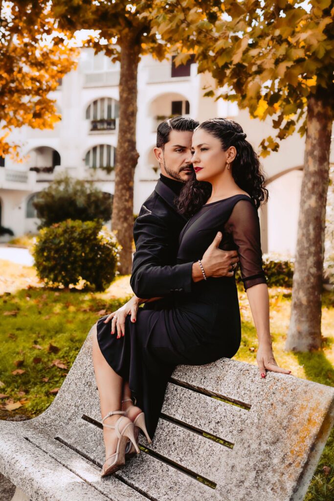 Un uomo e una donna vestiti di nero siedono vicini su una panchina di pietra in un parco con alberi autunnali e un edificio bianco sullo sfondo.