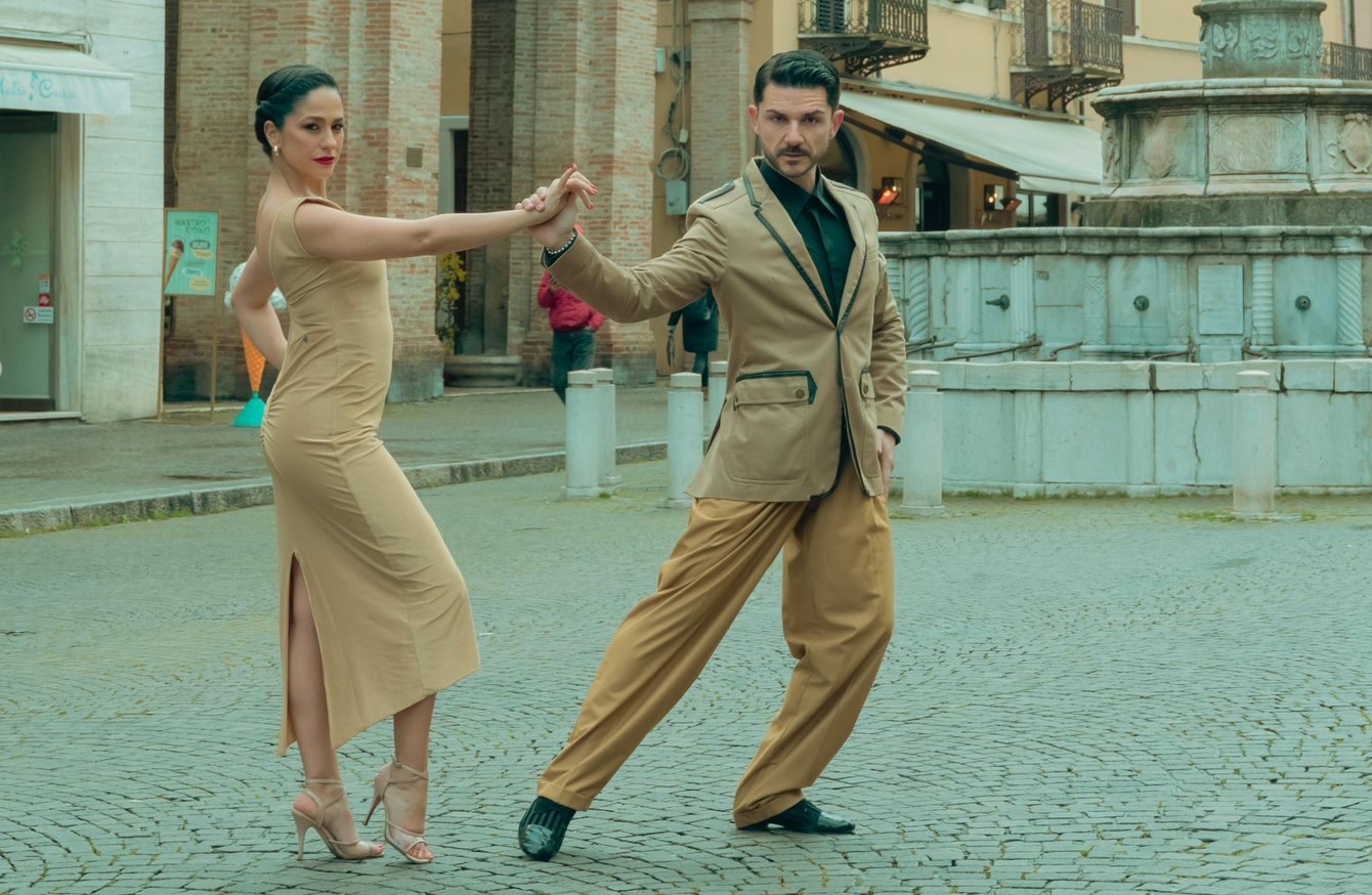 Un uomo e una donna in abiti abbronzati eseguono una posa di danza su una strada acciottolata di fronte a edifici storici e a una fontana di pietra.