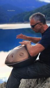 Un uomo con gli occhiali da sole siede in riva a un lago, suonando un tamburo a mano sulle ginocchia, con un paesaggio montano sullo sfondo.