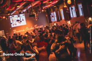 Un folto gruppo di persone balla il tango in una sala illuminata da luci calde, con la scritta “Buena Onda Tango” proiettata sulla parete.